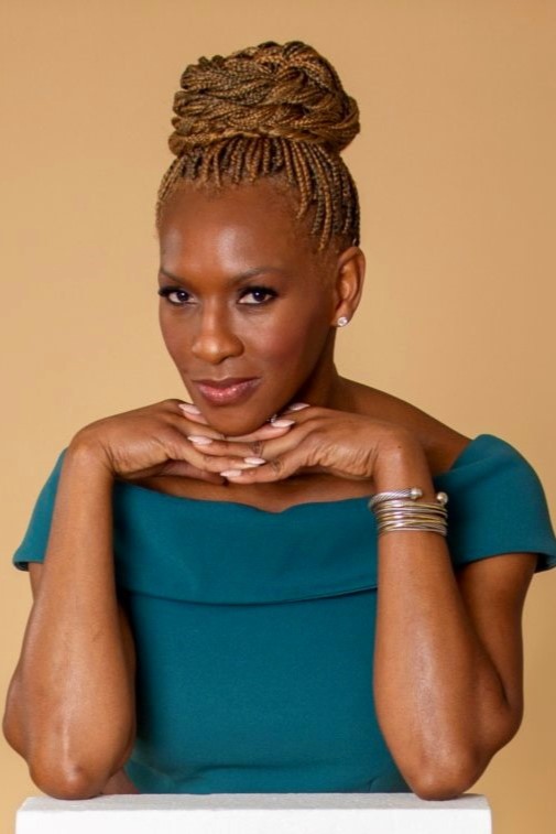 Black woman with teal top with elbows on top of a white box and interlocked fingers under her chin. There is a tan background.
