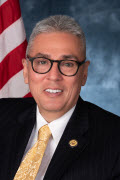 Andrés Gallegos, a hispanic male with gray hair wearing dark glasses and suit with white shirt and yellow tie, and an NCD lapel pin. Behind him is a sky background and U.S. flag.