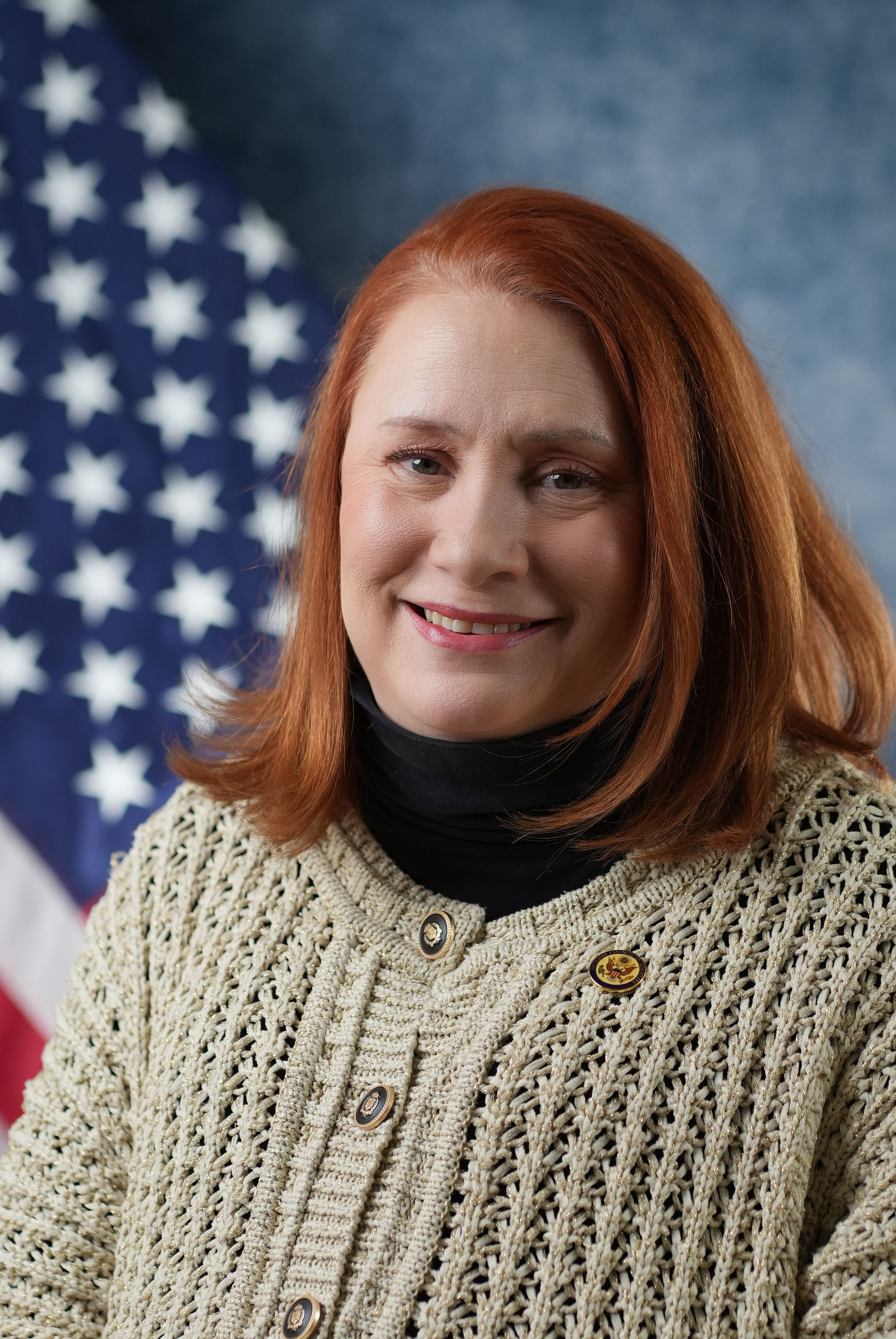 Kimberly Hill Ridley smiling. She has red hair and is wearing a beige sweater with black and gold buttons over black turtleneck shirt and NCD lapel pin.
